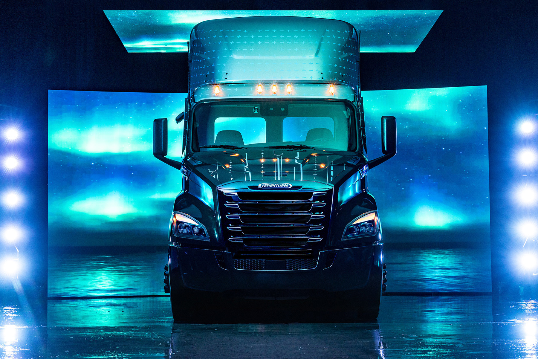 Freightliner semi-truck, forward facing, with headlights and roof lights on.